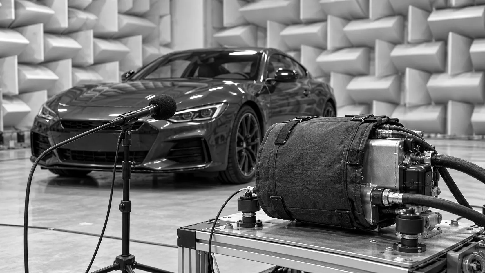 A compressor wrapped in a black acoustic jacket on a test bench in a semi-anechoic chamber, a microphone in front and a car behind