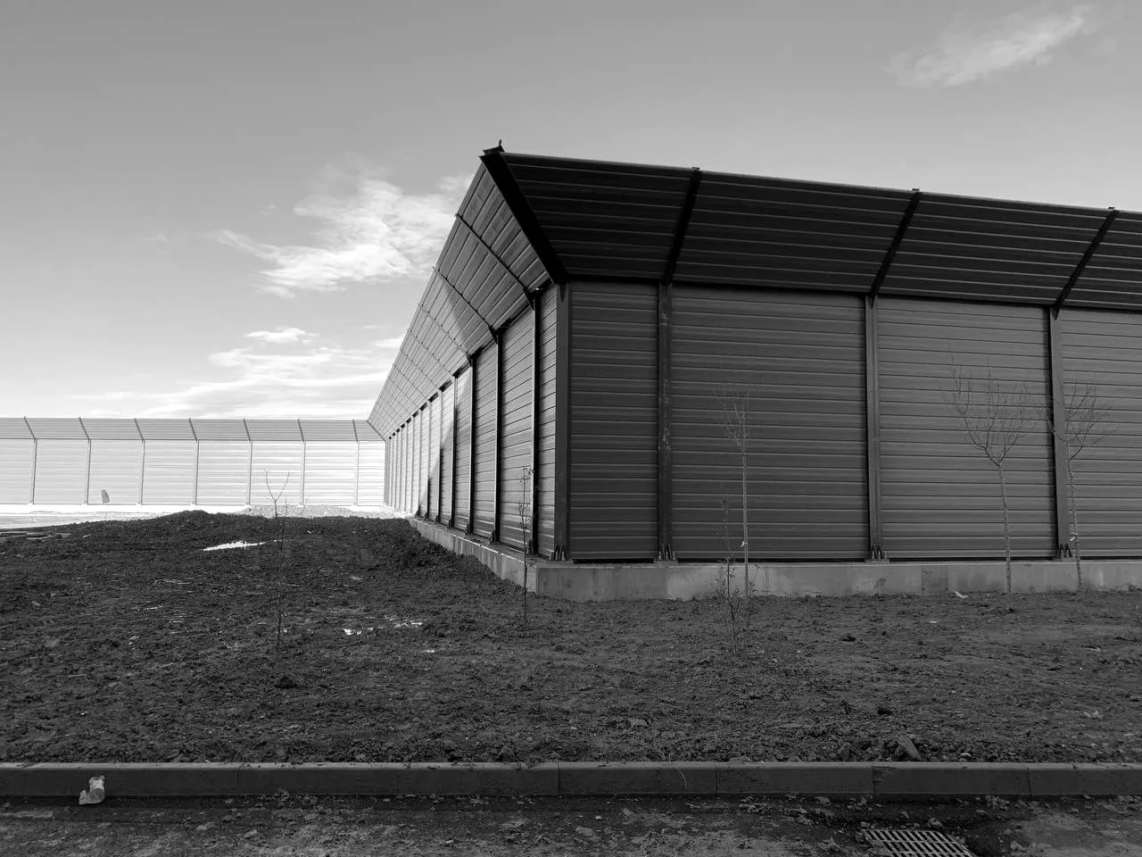 A long noise barrier with an angled top section, on a concrete base beside freshly levelled ground