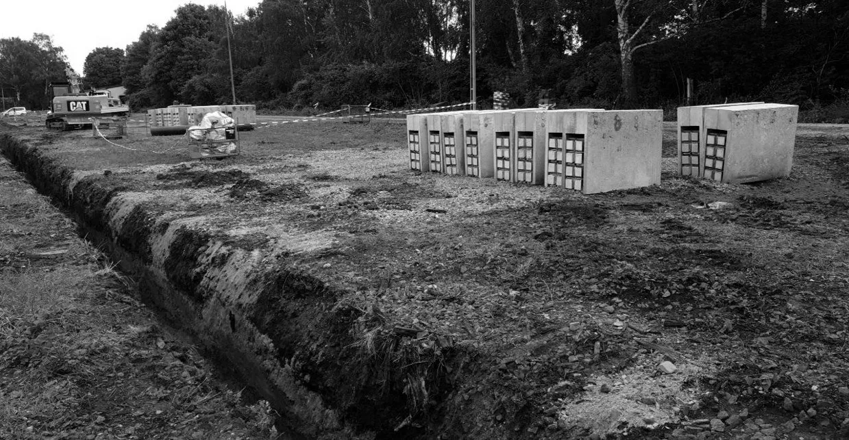 Concrete MetaVibration modules, their lattice cores showing, lined up beside an open trench, an excavator behind