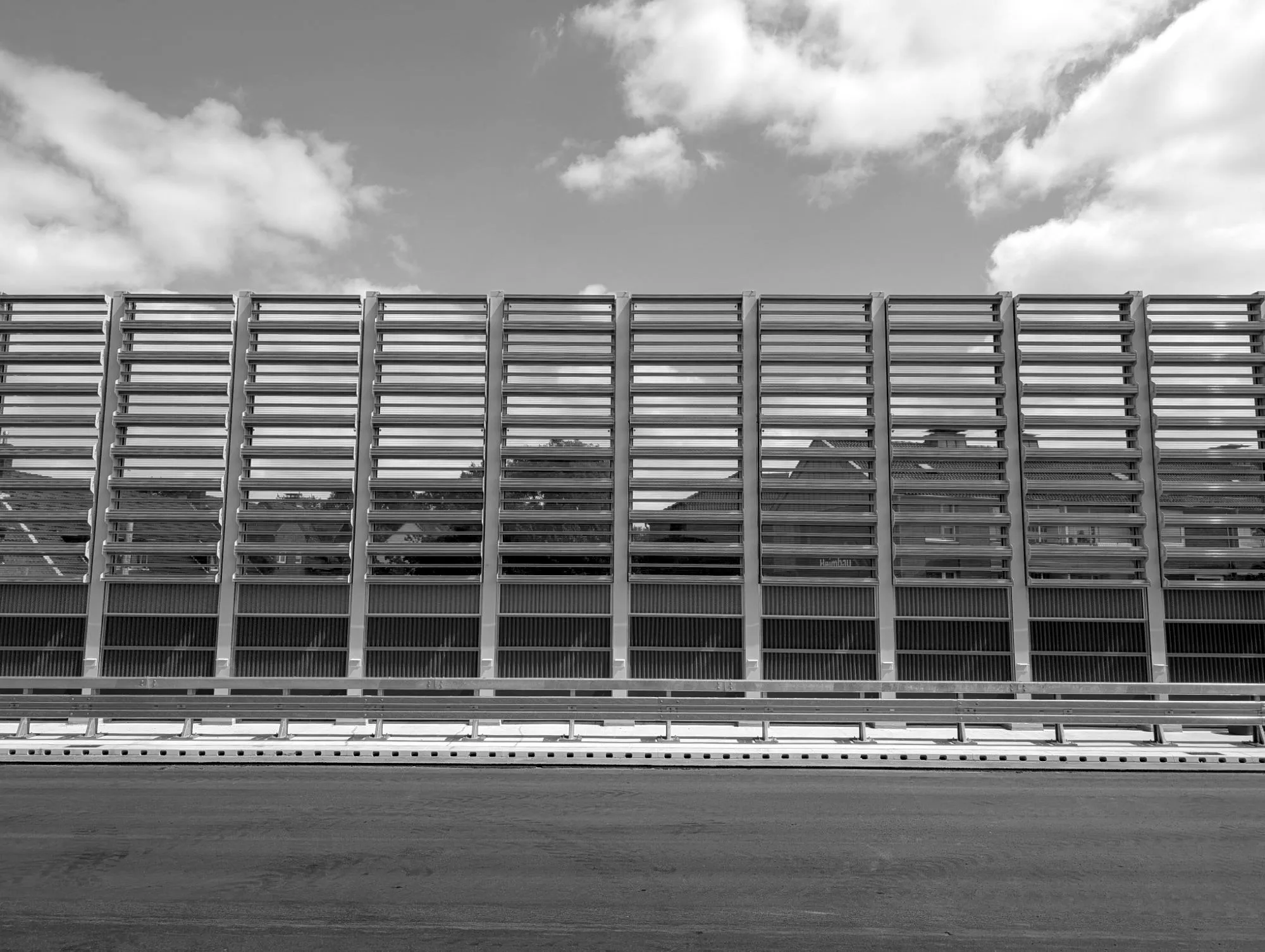 A transparent noise barrier of horizontal profiles along a new road, the roofs of houses visible through it